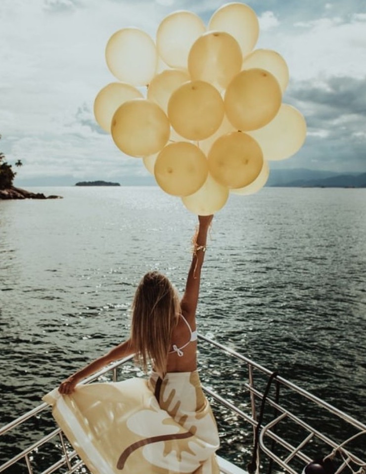 Woman holding golden balloons by the ocean, symbolising freedom and positivity, in an article on how to add joy to your day by Tess René Coaching.