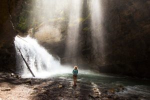 A person standing in front of a powerful waterfall, surrounded by sunlight streaming through the rocks, from an article about intentional rest by Tess Rene Coaching.