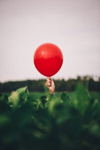 Hand holding a red balloon above green field, symbolising lightness and play — noted in Tess Rene Coaching’s joy writing