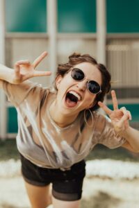 Young woman laughing with sunglasses, flashing peace signs, embodying spontaneous joy — highlighted in Tess Rene Coaching’s joy practices