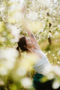 Woman stretching with arm overhead in sunlight, surrounded by greenery, symbolising release and vitality — shared in Tess Rene Coaching’s joy tools