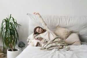 Woman stretching arms above her head in a soft sweater, relaxing in bed — from a Tess Rene Coaching feature on gentle somatic self-care