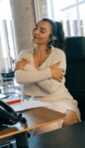 Woman hugging herself at desk, symbolising how writing your own paradigm brings clarity and reduces time spent being indecisive, in article by Tess René Coaching.