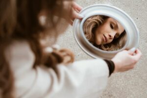 Woman looking into a mirror, featured in an article by Tess René Coaching on belonging versus fitting in