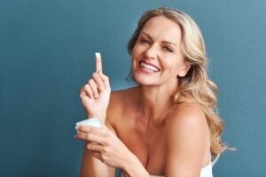 Smiling woman holding a small cream jar with finger raised, illustrating everyday self-care choices.