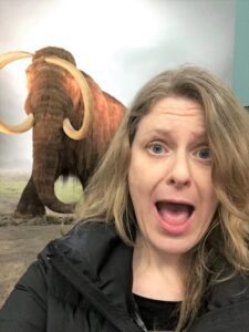 Surprised woman posing in front of a mammoth display, used in an article by Tess René Coaching about seeing intense emotions without judgment.