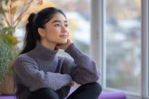A young woman sitting peacefully by the window, reflecting calmly after overcoming self-doubt in an article about facing fears by Tess René Coaching.