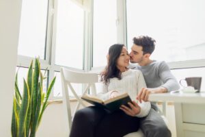 Couple reading together as he kisses her cheek, symbolising acceptance over admiration, in Tess René Coaching’s article.