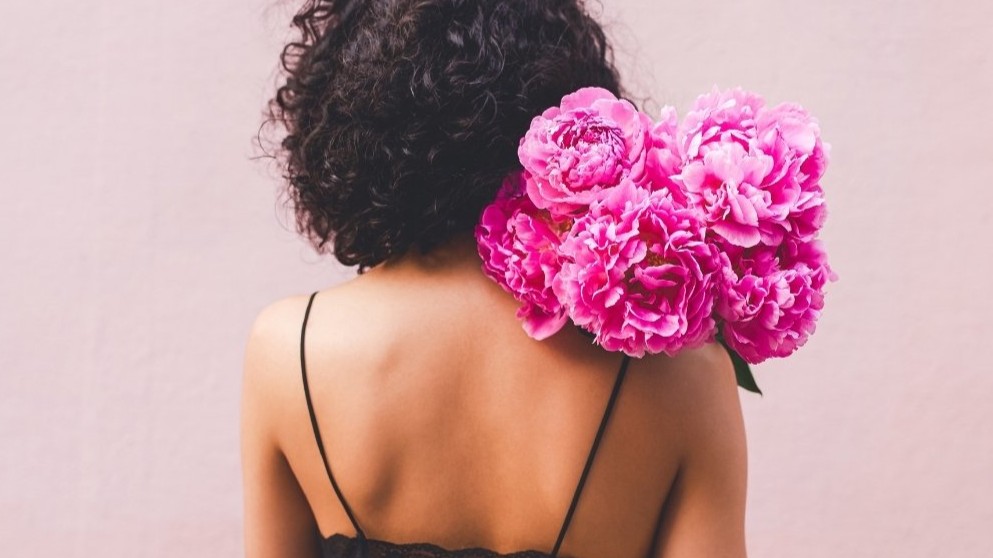 Back view of a woman holding pink peonies against a blush wall, symbolising self-kindness — featured image for “What Does Self-Compassion Really Mean?” by Tess René Coaching.