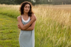 Woman standing in tall grass with eyes closed and arms around herself, reflecting calm self-compassion in this Tess René Coaching article on how to manage emotions.