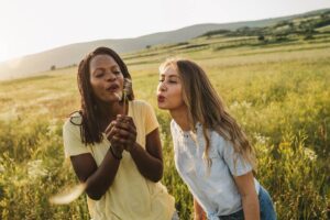Two friends blowing on a dandelion together in a sunlit field, symbolising opening my heart after disappointment, featured in an article by Tess René Coaching.