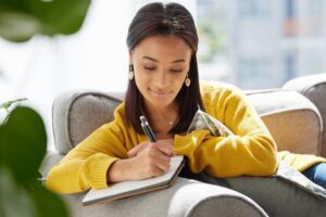Woman sitting on a sofa journaling and thinking, in an article by Tess René Coaching about calming perfectionism and anxiety.