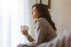 Woman looking out a window while holding a mug in a Tess Rene Coaching article about why hearts close