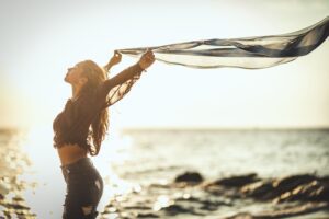 A woman holding a scarf at the shoreline at sunset, symbolizing personal agency in this Tess René Coaching article on dealing with flashbacks.
