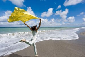 Woman running freely on the beach with a yellow scarf, featured in a Tess Renè Coaching article about how to live a happy life.