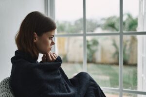 Woman wrapped in a dark blanket sitting by a window looking outside thoughtfully, featured in this Tess René Coaching article about the inner critic and self-doubt