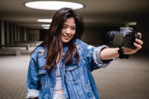 Young woman holding a camera and smiling while filming herself, featured in a Tess René Coaching article about healthy rituals.