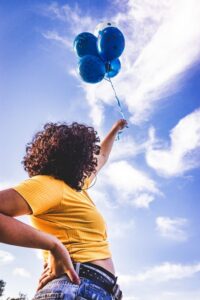a person stretching upward while holding a cluster of blue balloons, used in a Tess René Coaching article to represent interrupting old patterns and creating a new direction