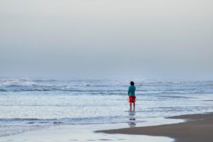 person standing alone in shallow ocean water facing the horizon under a grey sky in this Tess René Coaching article about releasing self punishment