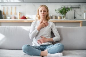 woman sitting cross legged on a couch with eyes closed and hands on chest and stomach in this article by Tess René Coaching about vulnerability and how to forgive yourself.
