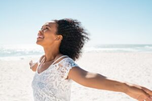 woman smiling with arms outstretched standing on a beach in sunlight in this Tess René Coaching article about freedom after self forgiveness