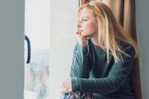 woman sitting by a window resting her chin on her hand and looking outside in this article by Tess René Coaching about guilt and self forgiveness