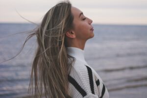 Woman with eyes closed breathing deeply beside ocean with wind moving through her hair in this Tess Renè Coaching article about how to enjoy life again and expanding emotional range