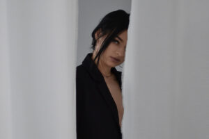 Dark-haired woman standing quietly between white curtains with a thoughtful expression in this Tess Renè Coaching article about how to feel better about myself and emotional wellbeing