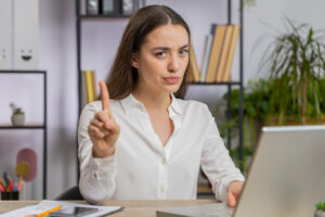 Woman raising one finger while sitting at a desk with a laptop in a Tess René Coaching article section about hiding your truth, speaking clearly, and building self trust