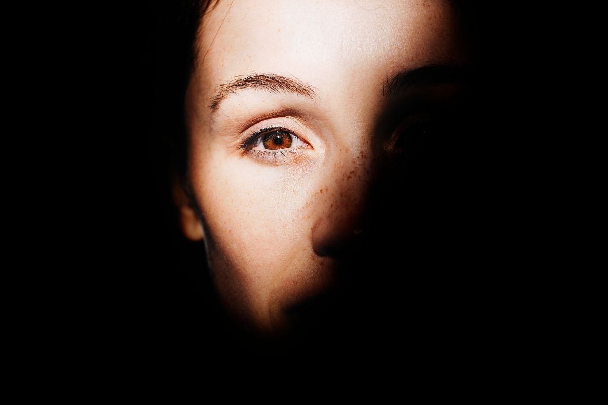 Close portrait of a woman’s face emerging from deep shadow, with one eye lit and looking forward, representing self doubt, emotional overwhelm, and the search for self connection.