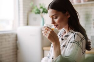 A woman standing in a bright kitchen with her eyes closed as she smells a warm drink, representing calm, pleasure, presence, and small moments of joy.