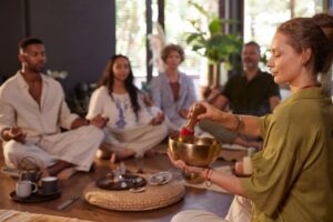 A woman playing a singing bowl while five people sit together in a relaxed circle, representing safe connection, shared presence, and nervous system regulation.