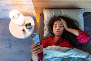 Woman lying awake in bed at night looking at her phone, appearing tired and worried, in a Tess René Coaching article section about why we feel worse when limiting beliefs and negative self talk go unchallenged
